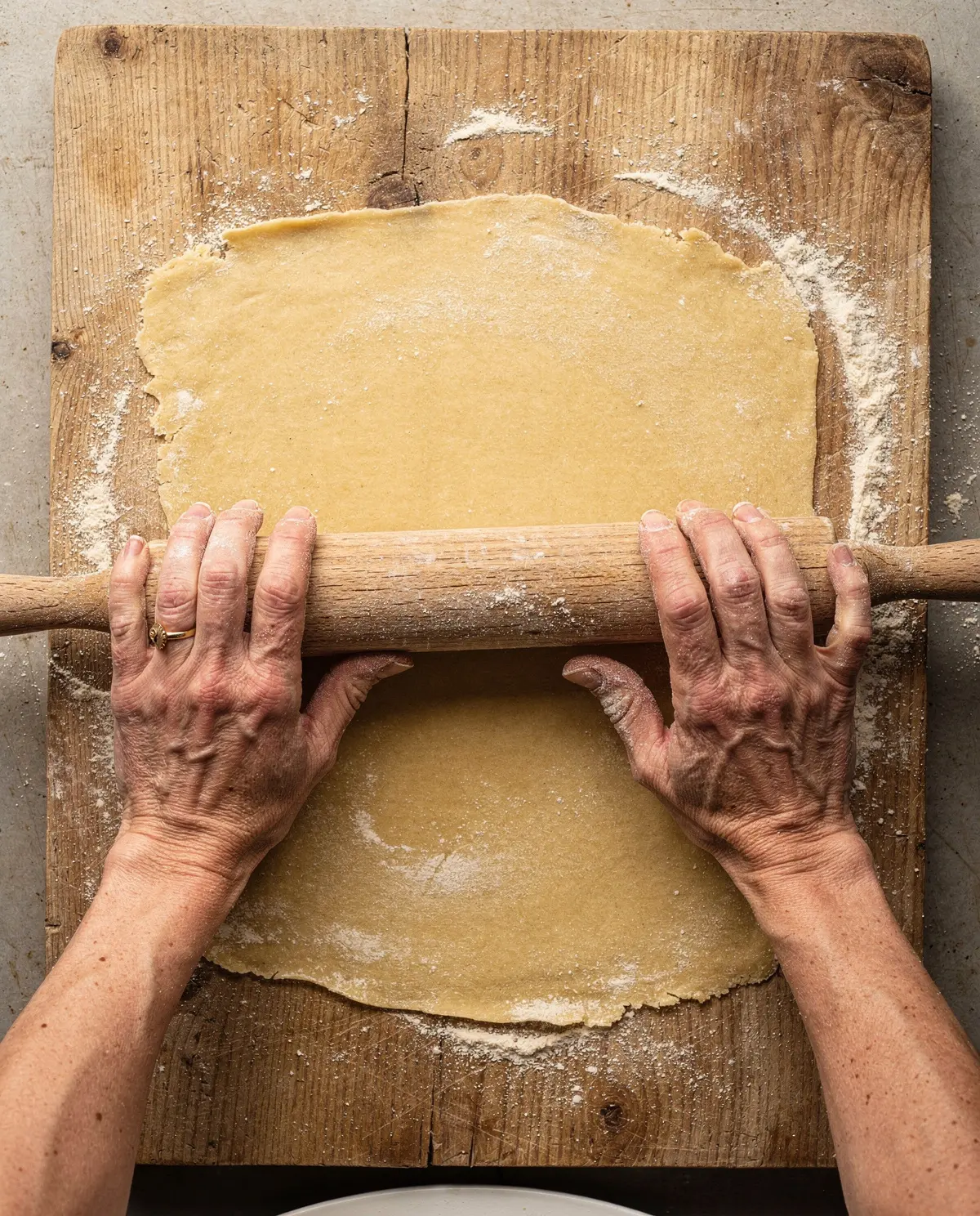 Hands rolling a sheet of pasta dough with a worn wooden pin